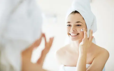 Una mujer sonriente con toalla en la cabeza se aplica crema en la cara, un paso esencial para como cuidar la piel de la cara.