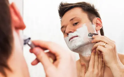 Hombre con espuma de afeitar en la cara, afeitándose con una maquinilla. ¿Es mejor afeitarse antes o después de la ducha?
