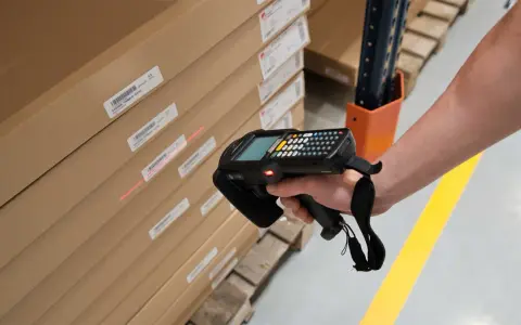 Mano escaneando cajas apiladas con un dispositivo, usando la **fórmula de Wilson** para optimizar el inventario.