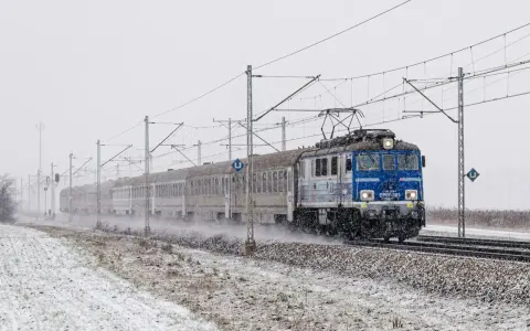 Niebieska lokomotywa z pociągiem na tle zaśnieżonego krajobrazu i słupów trakcyjnych.