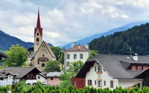 Un borgo pittoresco in Val d'Isarco, con una chiesa dal campanile rosso svettante tra case tradizionali e montagne boscose.