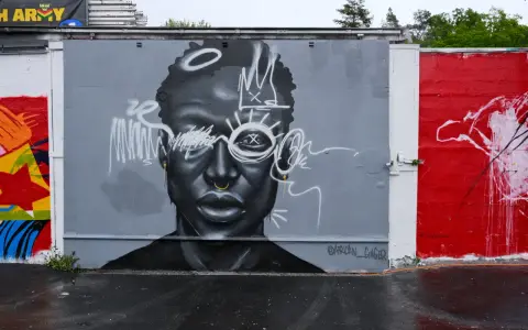 En imponerande **graffiti** av ett ansikte med en krona och solstrålar runt ögonen. Konstverket är målat i gråskala med vita detaljer.