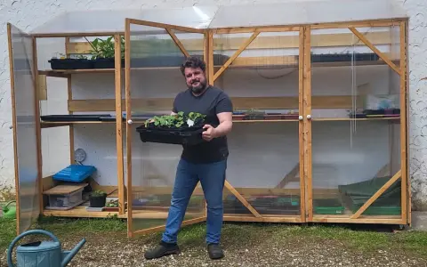 Un homme souriant tient un plateau de jeunes plants devant sa serre pour semis fait maison, remplie d'étagères de semis.