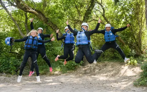 Cinco personas, incluyendo niños, saltan alegres con trajes de neopreno y chalecos salvavidas en un entorno natural, listos para una aventura en Huesca.