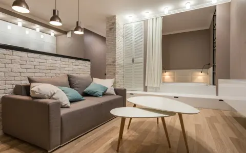 Nowoczesny salon z sofą i stolikami kawowymi. W tle widoczna ściana ze starej cegły, łóżko i szafa.