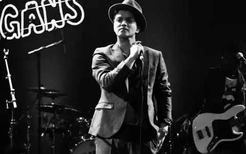 Bruno Mars i Sverige, med hatt och kavaj, sjunger i mikrofonen. Bakom honom syns ett band och neonljus.
