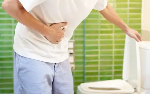 Mężczyzna trzymający się za brzuch, stojący obok otwartej muszli klozetowej.