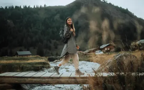 Kobieta w stylu gorpcore przemierza drewniany mostek nad strumieniem, otoczona górami i lasem.
