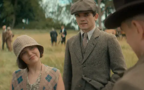 Giovane donna con cappello a scacchi e gilet a rombi, accanto a un uomo con berretto e giacca di tweed. Sullo sfondo, i sette quadranti di un campo.