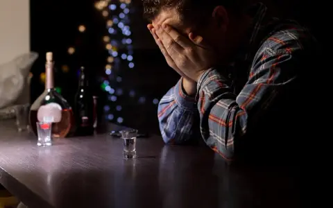 Mężczyzna z rękami na twarzy przy barze, obok butelka i kieliszek. Obraz symbolizuje przymusowe leczenie alkoholika i jego walkę.