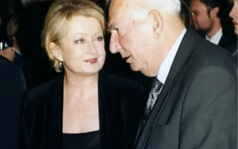 Ewa Holoubek w czarnej sukni i marynarce rozmawia z mężczyzną w garniturze.