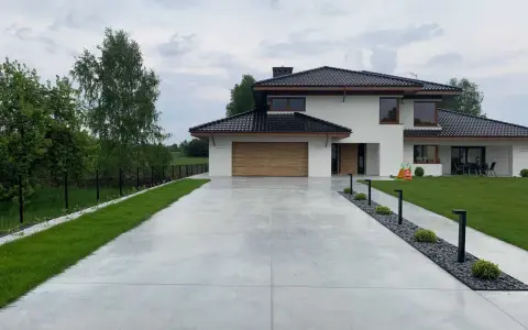 Nowoczesny dom z drewnianą bramą garażową, otoczony zielenią. Szeroki podjazd betonowy prowadzi do wejścia, a wzdłuż niego biegną lampy i rabaty z kamieniami.