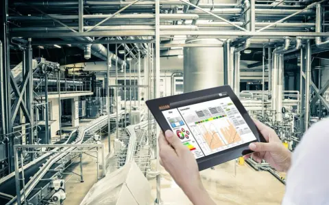Operator kontroluje linię produkcyjną za pomocą tabletu, na którym widnieje wykres zużycia energii. W tle widać skomplikowany system rur i taśmociągów. Czujnik podczerwieni może być kluczowy dla monitorowania procesów.