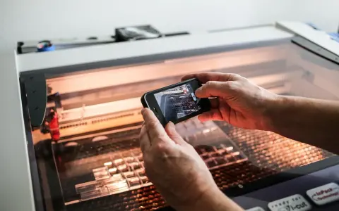 Hands hold a phone, capturing a laser cutter working on intricate acrylic projects.