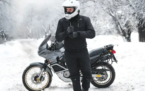 Motociclista con casco bianco e abbigliamento invernale nero, pronto per un'avventura in moto inverno su una strada innevata.