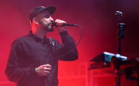 Wokalista z brodą i czapką na głowie śpiewa do mikrofonu na scenie. Czerwone światła tworzą atmosferę koncertu. Tauron Nowa Muzyka line-up.