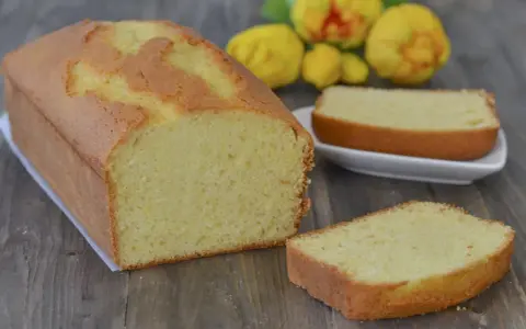 Un soffice plumcake al limone, appena sfornato, con una fetta su un piattino bianco e fiori gialli sullo sfondo.