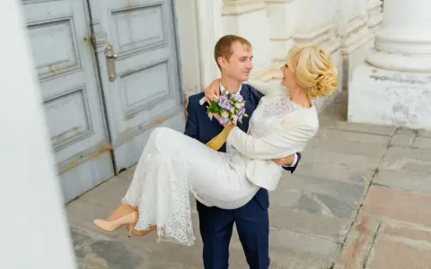Pan młody niesie pannę młodą na rękach. Zastanawiasz się nad skutkami spóźnienia się z rezerwacją fotografa? Dlaczego 18 miesięcy to minimum?