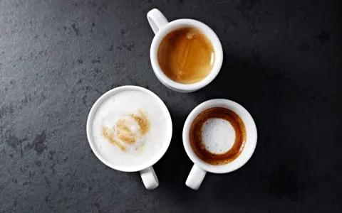 Trzy białe filiżanki z kawą: espresso, latte macchiato i kawą z pianką na ciemnym tle.