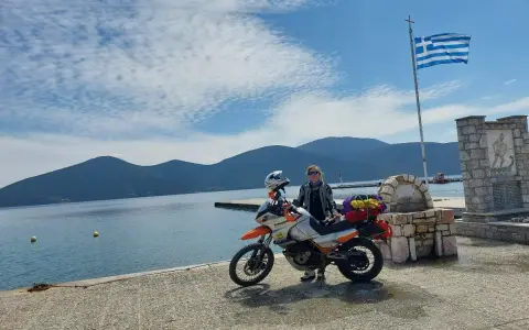 Podróżniczka z motocyklem nad grecką wodą, pod flagą Grecji. Czy Grecja jest droga? Ta podróż wygląda na wartą każdej ceny!