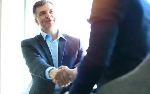 Uśmiechnięty mężczyzna w garniturze ściska dłoń partnerowi biznesowemu. To obrazuje kluczowe zadania specjalisty ds. sprzedaży: budowanie relacji i finalizowanie transakcji.