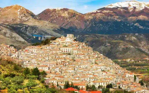 Un pittoresco borgo calabrese si aggrappa a una collina, con case in pietra e tetti rossi che si estendono fino alle montagne innevate.