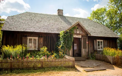 Drewniana apteka w Muzeum Wsi Kieleckiej, otoczona kwitnącym ogrodem i ogrodzona płotkiem.