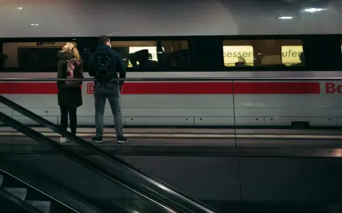 Para stojąca na peronie przed pociągiem, obok schodów ruchomych.