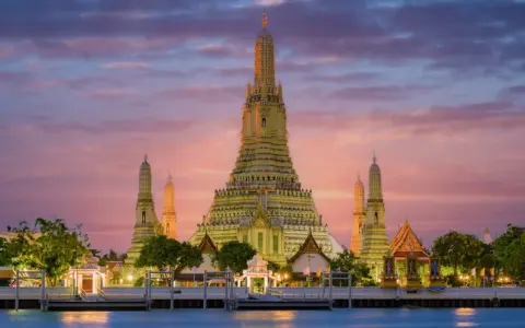 Il **tempio dell'alba Bangkok** (Wat Arun) si erge maestoso al tramonto, con le sue guglie dorate che brillano contro un cielo rosa e viola.