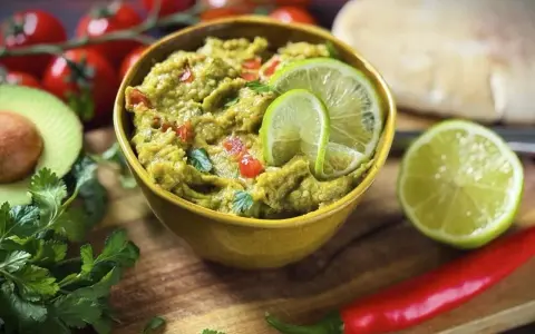 Frisches Guacamole-Rezept in einer gelben Schüssel, garniert mit Limettenscheiben. Daneben liegen Avocado, Koriander, Tomaten und Chili.