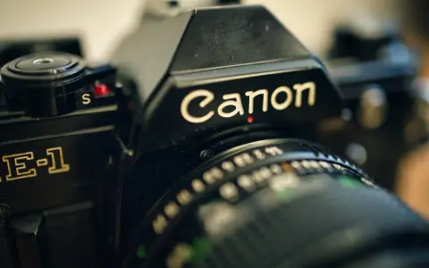 Zbliżenie na czarną lustrzankę Canon AE-1. Widoczny napis "Canon" i fragment obiektywu. Idealny do połączenia z aparatem.