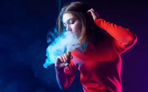 Joven exhala humo de un vaper, rodeada de luces azules y moradas.