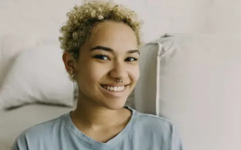 Uśmiechnięta młoda kobieta z kręconymi blond włosami i kolczykiem w nosie (septum piercing).