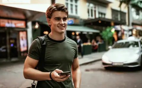 Ung man med mobiltelefon i handen, på en stadsgata med bilar och butiker. Känns som vårt hem utomlands.