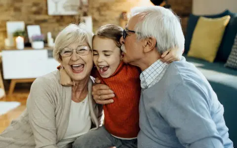 Una niña ríe mientras su abuelo le da un beso y su abuela la abraza. Así de felices los abuelos son.
