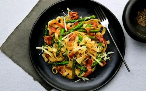 Pyszny makaron ze szparagami, chrupiącym boczkiem i parmezanem na czarnym talerzu. Idealny na wiosenny obiad.