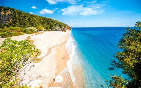 En orörd albanien strand med turkost vatten, vit sand och gröna kullar. En liten flod rinner ut i havet.