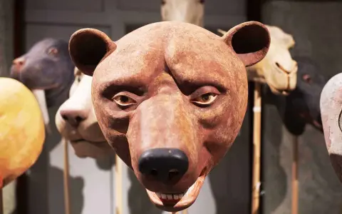 En brun björnmans ansikte, gjord av papier mache lim, med andra djurmasker i bakgrunden.