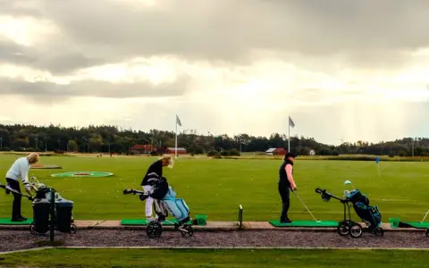 Kvinnor tränar golf på en driving range i Sverige. Solstrålar bryter igenom molnen.