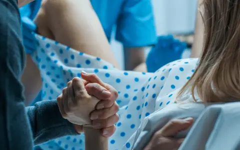 Wsparcie bliskiej osoby podczas porodu. Dłoń partnera trzyma dłoń rodzącej, która ma już widoczny czop śluzowy.