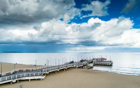 Długie molo w Brzeźnie wchodzi w spokojne morze. Na piaszczystej plaży widać kilka osób i samochody na molo.