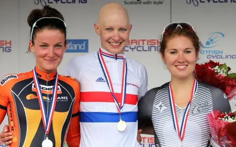 Joanna Rowsell, a bald cyclist in a white and red jersey, smiles on the podium with two other medalists.