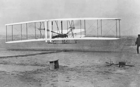 Wright Flyer mit Doppelruder hebt ab, Pilot liegt auf dem Flügel. Ein Mann beobachtet den historischen Flug.