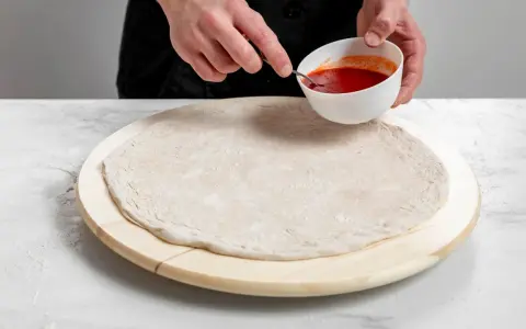 Przygotowanie ciasta na pizzę bez drożdży. Dłonie nabierają sos pomidorowy z miseczki, by rozprowadzić go na okrągłym spodzie.