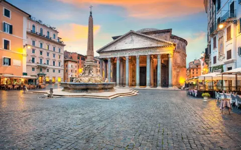 Piazza illuminata con il Pantheon, una chiesa maestosa, e una fontana con obelisco. Tavolini all'aperto invitano a godersi la sera romana.