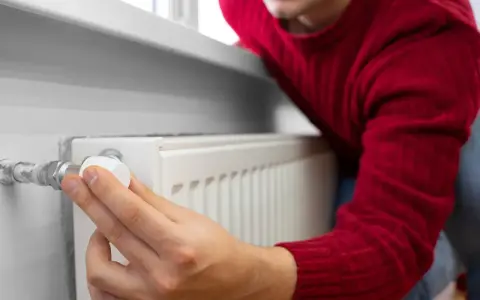 Mężczyzna w czerwonym swetrze reguluje termostat grzejnika, oszczędzając energię. To jego sposób na wykorzystanie bonu ciepłowniczy.