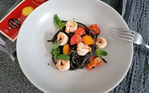 Czarny makaron z krewetkami, pomidorkami i bazylią na talerzu. Obok widoczna paczka spaghetti nero di seppia.