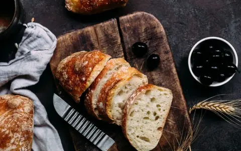 Frisches Brot, aufgeschnitten auf einem Holzbrett, daneben Oliven. Perfekt für den Gasgrill bei richtiger Temperatur.