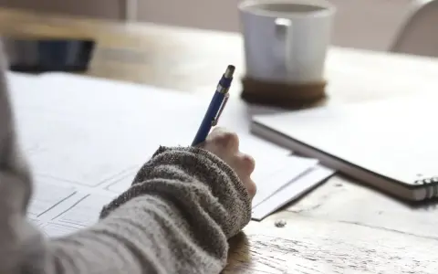 Ręka pisze długopisem na papierze, planując, jak napisać książkę. Obok kubek kawy i notatnik.