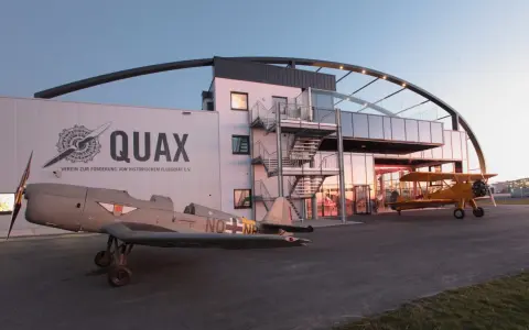 Zwei historische Flugzeuge stehen vor dem Quax Hangar. Ein graues Propellerflugzeug im Vordergrund, ein gelbes Doppeldeckerflugzeug daneben.
